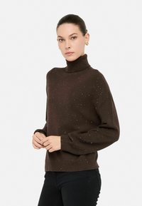 Vrouw in een donkerbruin gemêleerd coltrui en zwarte broek, met haar opgestoken en gouden hartvormige oorbellen.