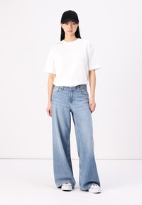 T-shirt bianco a maniche corte abbinato a jeans larghi blu chiaro e sneakers bianche, accessoriato con un berretto nero. Tessuto morbido e stile casual.