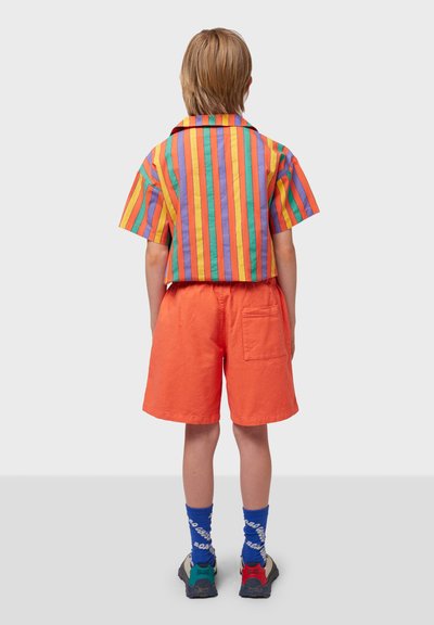 Gestreifte Kurzarmbluse mit orangefarbenen, grünen, lila und blauen Streifen, kombiniert mit orangefarbenen Shorts und blauen gemusterten Socken und Schuhen.