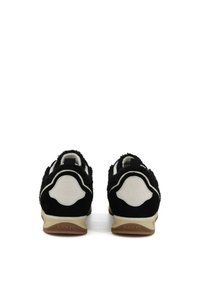 Une paire de baskets noir et blanc avec un tissu texturé et "BACK 70" sur des semelles beige, montrée de l'arrière sur un fond blanc.