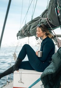 Frau in einem dunkelgrünen Thermanzug sitzt auf einem Segelboot und hält einen Metallbecher. Sie trägt schwarze knielange Socken, und um sie herum ist Segelausrüstung zu sehen.
