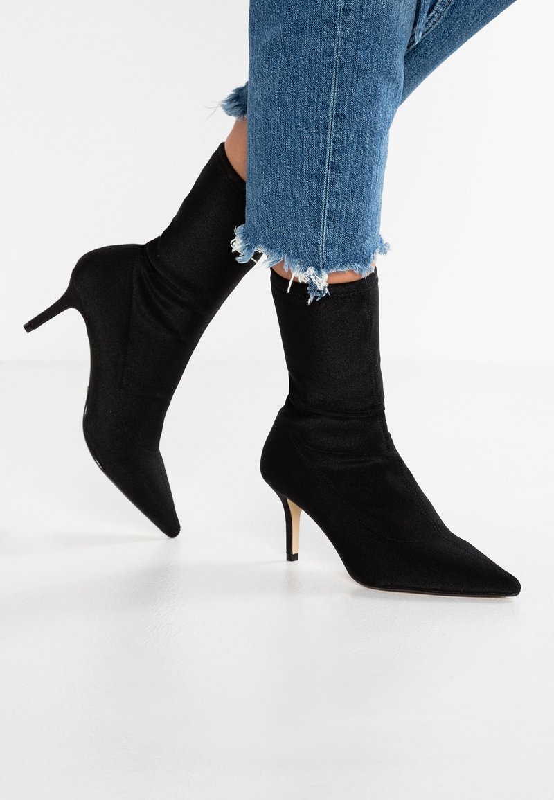 Femme portant des bottines noires à bout pointu et talons hauts, associées à un jean bleu à ourlet effiloché, sur fond blanc.