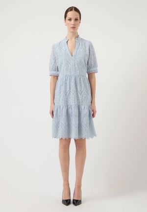 Femeie în picioare, purtând o rochie bleu deschis din dantelă cu ochiuri, cu mâneci scurte bufante și pantofi cu toc ascuțit negri, pe un fundal alb simplu.