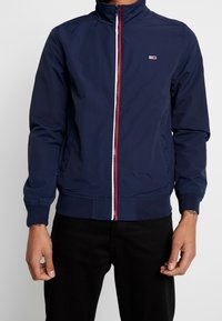 Marineblaue Bomberjacke aus leichtem Material mit hohem Kragen, ausgestattet mit einem roten und weißen Reißverschluss sowie gerippten Bündchen und Saum.