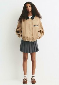 Jeune femme aux cheveux bouclés portant une veste en velours côtelé beige, une jupe plissée grise, des chaussettes blanches et des baskets marron, se tenant devant un mur uni.