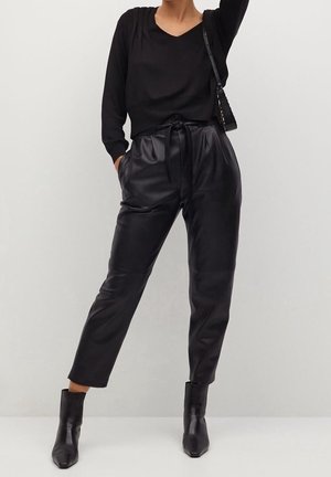 Pull noir à col en V associé à un pantalon en cuir noir taille haute et ajusté. Des bottines noires montant à la cheville et un petit sac à main noir complètent la tenue.