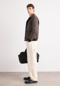 Bruine, textuurjasje, crèmekleurige wijde broek en zwarte schoenen. Model houdt een zwarte tas vast. Eenvoudig, modern ontwerp met contrasterende kleuren.