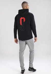 Schwarzer Kapuzenpullover mit einem großen roten und grauen "9"-Grafik auf dem Rücken, kombiniert mit hellgrauen Jogginghosen und schwarzen Schuhen.