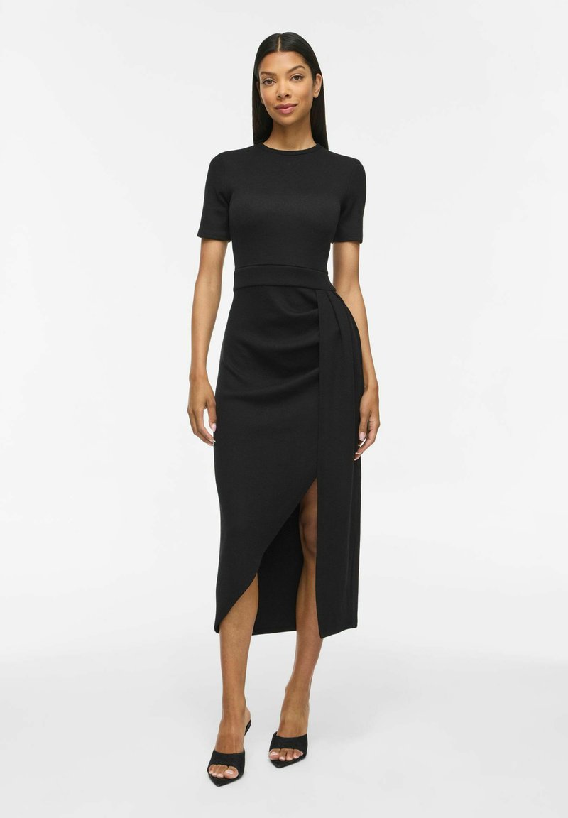 Femme debout face à l’objectif, portant une robe noire ajustée à manches courtes et ourlet asymétrique, ainsi que des talons noirs ouverts, sur fond uni.