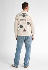 Chaqueta de algodón de color claro con varios parches bordados en la parte posterior, combinada con unos vaqueros de mezclilla azul claro de corte holgado.