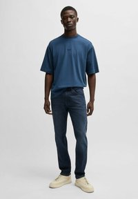 Blå kortärmad skjorta med vertikala randiga detaljer, kombinerad med mörka jeans och ljusa sneakers. Enkel design, avslappnad passform.