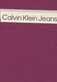Lila Baumwollstoff mit einem Etikett, auf dem "Calvin Klein Jeans" in fettem schwarzen Text steht, ergänzt durch einen kontrastierenden grauen Streifen.