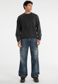 Kool zwarte crewneck trui met logo, gecombineerd met losse blauwe cargobroeken met meerdere zakken. Bruine veterschoenen maken de outfit compleet.