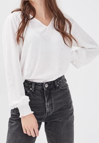 Femme portant un blouse blanche à manches longues avec de subtils pois floraux et des poignets smockés, associée à un jean noir décoloré taille haute.