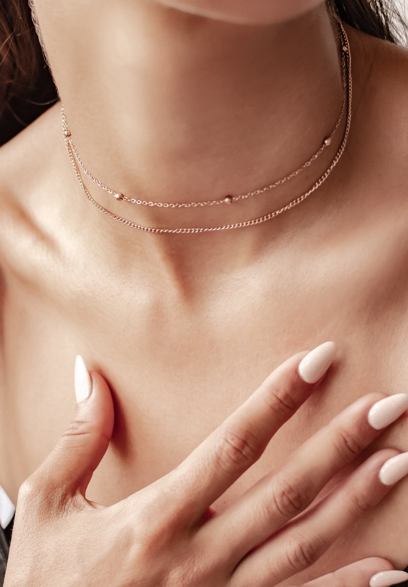 Dwie delikatne, warstwowe chokery wykonane z różowego złota, ozdobione małymi okrągłymi koralikami i cienkimi łańcuszkami, spoczywające na gładkiej szyi.