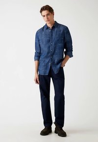 Chemise bleue à manches longues avec un motif, col à boutons et poche poitrine. Associée à un pantalon sombre et des chaussures marron.