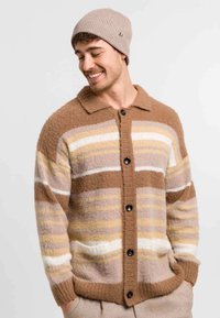 Young man wearing a brown, beige, and white striped buttoned cardigan and a light brown knit beanie, smiling with hands in pockets.