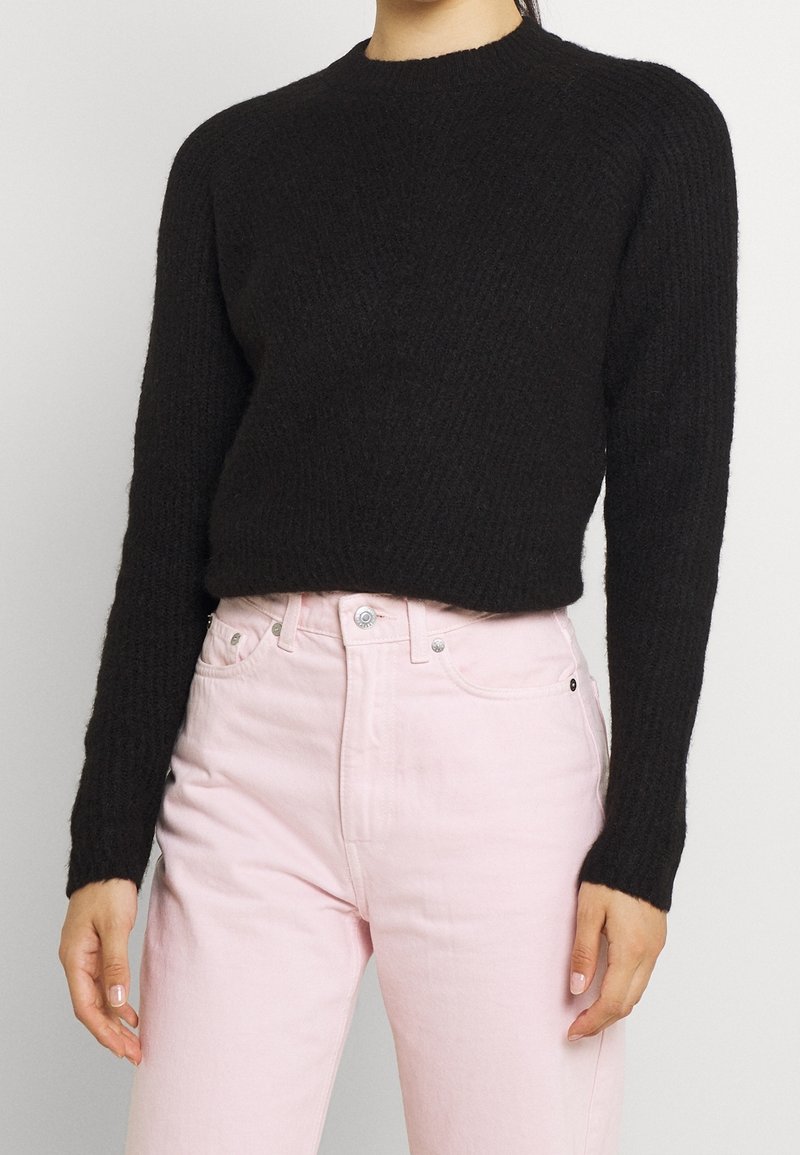 Suéter negro de canalé con cuello redondo y mangas largas, combinado con jeans rosa claro de talle alto. Textura suave y estilo ajustado.