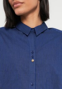 Camicia a righe blu navy con scollo a collo, tre bottoni e un bottone decorativo dorato. Presenta una tasca sul petto a sinistra.