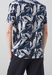 Košile s krátkým rukávem vyrobená z lehkého materiálu; má tmavě modré pozadí se vzorem listů v bílé a modrých odstínech.