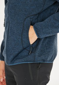 Laikapuku su neperšlampamu paviršiumi, turintis šonines kišenes ir kontrastingus juodus užtrauktukus. Derinamas su juodais kelnais, kuriuose yra užtrauktukai.