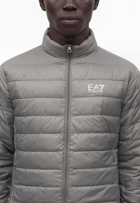 Άνδρας που φορά γκρι καπιτονέ μπουφάν EA7 Emporio Armani με φερμουάρ μπροστά, εμφανίζεται από το μέσο του στήθους μέχρι το πηγούνι σε λευκό φόντο.