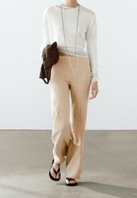 Lichtgrijs shirt met lange mouwen, beige gestreepte broek met wijde pijpen, zwarte teenslippers, met een getextureerde donkerbruine tas, tegen een effen achtergrond.