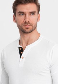 Weißes Langarm-Henley-Shirt mit schwarzem Knopfleiste und drei Knöpfen. Glatte Textur, taillierte Passform, ohne Muster oder Akzente.
