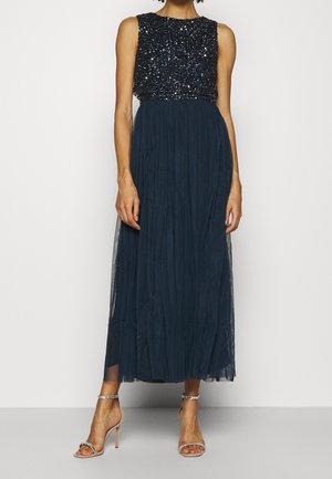 Abito lungo con gonna plissettata blu navy e corpetto di paillettes. Presenta maniche a mantella e una vita aderente, completato da tacchi con cinturino alla caviglia.