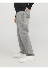 Osoba v světle šedých rovně střižených jeans a bílých teniskách, s jednou rukou v kapse černé mikiny.