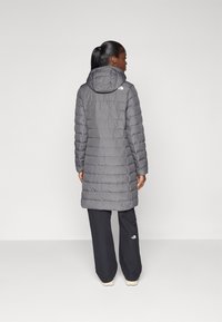 Pelēks puffers ar kapuci, ar quilted dizainu un garāku griezumu. The North Face logotips redzams augšējā mugurā.