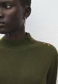 Pull en maille vert olive avec un col côtelé haut et des boutons ronds décoratifs le long d'une épaule ; associé à une boucle d'oreille en or.