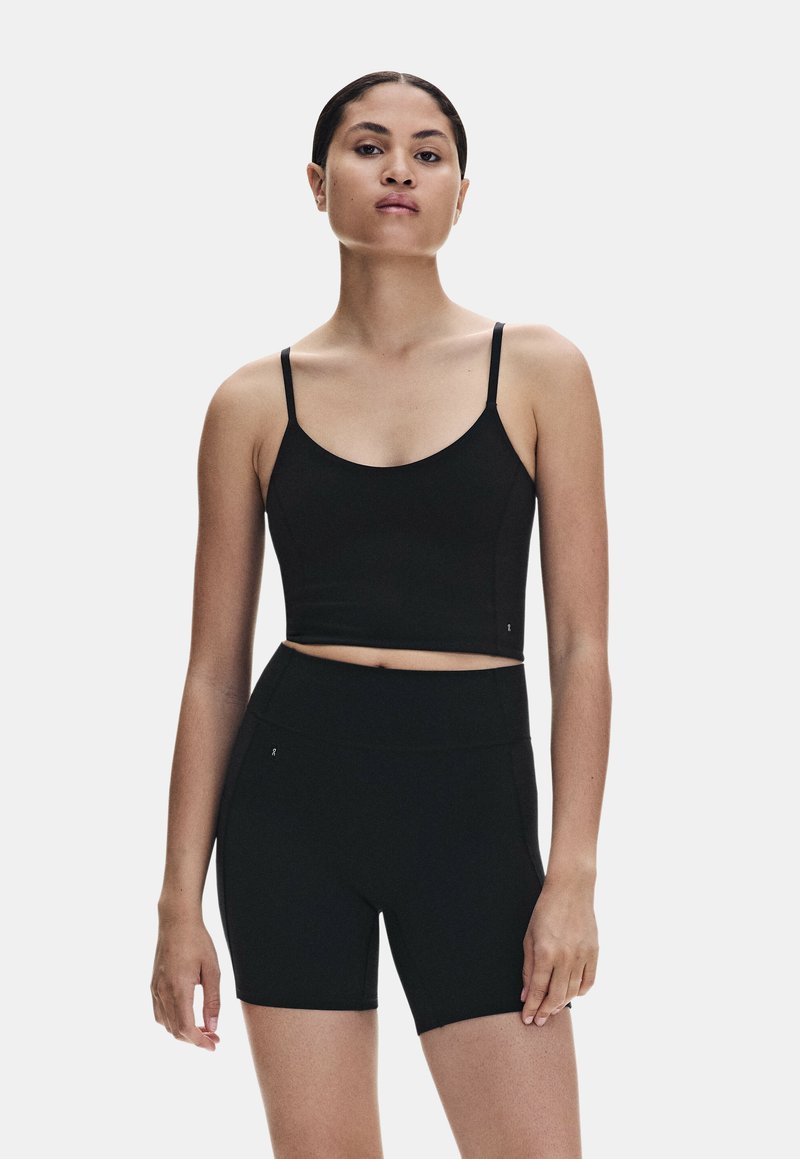 Femme aux cheveux attachés portant un crop top noir à fines bretelles et un short cycliste noir taille haute sur un fond uni.