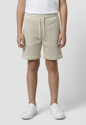 Dijete u bež mrežastim pletenim kratkim hlačama s vezicom u struku, bijelim tenisicama i bijelom majicom stoji ispred jednobojne pozadine.