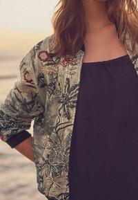 Nainen, joka wearing glitterikukkakuosiset bomber-takki mustan topin päällä, seisoo ulkona käsi lantiolla.