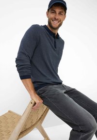 Polo a maniche lunghe navy testurizzato con colletto scuro, abbinato a pantaloni in denim grigi. Il modello è seduto su una sedia di legno con seduta in tessuto intrecciato.