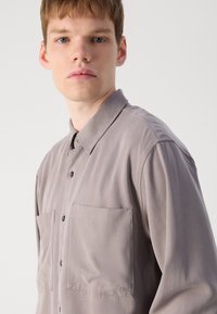 Šviesiai pilkas marškinis su apykakle, pagamintas iš lygaus audinio, turintis dvi krūtinės kišenes, sagų užsegimą ir siuvinėtą logotipą ant apvado.
