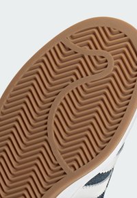 Kumipohjallinen lenkkari, jossa on aaltoileva herringbone-kuvio vaaleanruskeana ja tekstuuripainatuksilla. Suorat valkoiset reunat korostavat muotoilua.