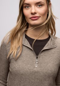 Ljust brun ribbad zip-up tröja med en silverfärgad dragkedja och rund dragkedjeknop, med hög krage och lager-på-lager över en mörkbrun skjorta.