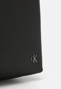 Black leather bag featuring a smooth texture and a small, shiny embossed "CK" logo on the front. Simple design with a rectangular shape.