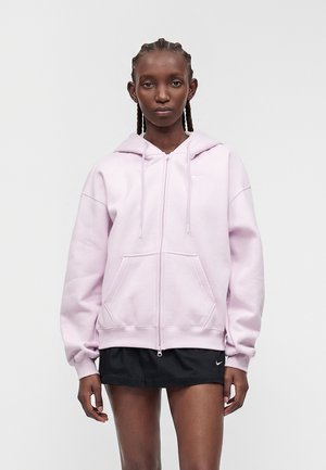 Femme aux cheveux tressés portant un sweat à capuche zippé rose clair et un short Nike noir, debout sur un fond clair uni.