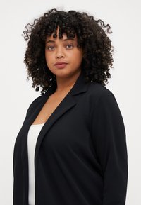 Černý blazer s hladkou texturou, otevřeným předním dílem a klopami, nošený přes bílý top. Modelka má kudrnaté vlasy a neutrální líčení.