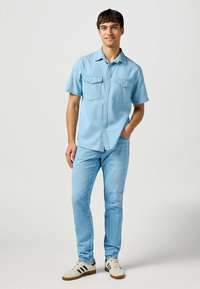Camicia azzurra a maniche corte con due tasche sul petto, chiusura con bottoni, abbinata a jeans azzurri e sneakers beige con strisce nere.