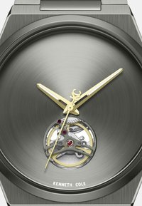 Grijze metalen horlogekast met gouden wijzers. Bevat een transparante sectie die de tandwielen en robijnaccenten onthult. Geborsteld metalen kast.