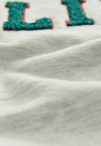 Vit tyg med lätt textur och veck, prytt med gröna och röda broderade bokstäver. Fokusera på materialet och detaljerna i broderiet.