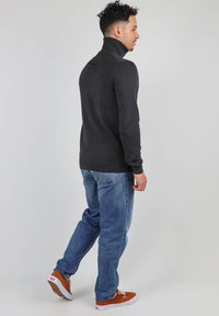 Pull en col roulé gris foncé, texture douce, coupe ajustée. Pantalon en jean bleu et chaussures en daim marron, vu de dos.