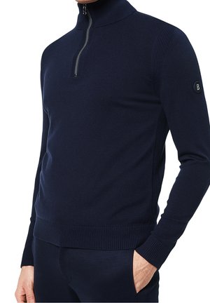 Hombre vistiendo un suéter azul marino de cremallera cuartilla con puños y dobladillo acanalados, mano izquierda en el bolsillo, y parche de marca en la parte superior izquierda de la manga.
