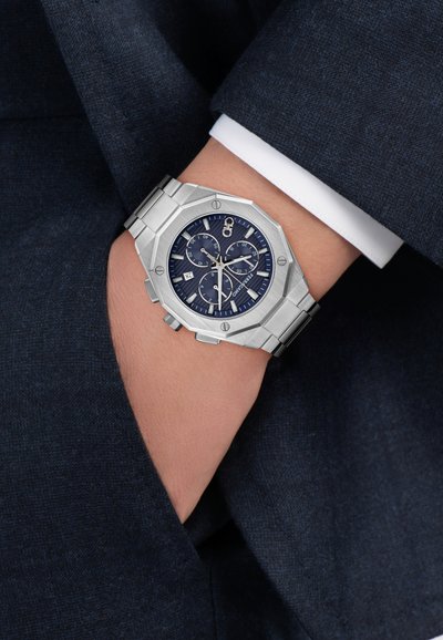 Ezüstszínű nyolcszögletű óra kék számlappal, rozsdamentes acél szíjjal, kronográf funkciókkal és dátumkijelzéssel, sötét öltönyös csuklón viselve.