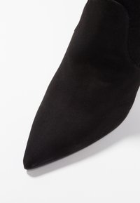 Stivaletto alla caviglia in suede nero con punta affilata e una texture liscia, dotato di pannelli laterali elastici per una silhouette aderente.