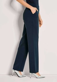Vrouw in navyblauwe wijde broek en zilveren hoge hakken, hand in de zak, staand tegen een effen achtergrond.